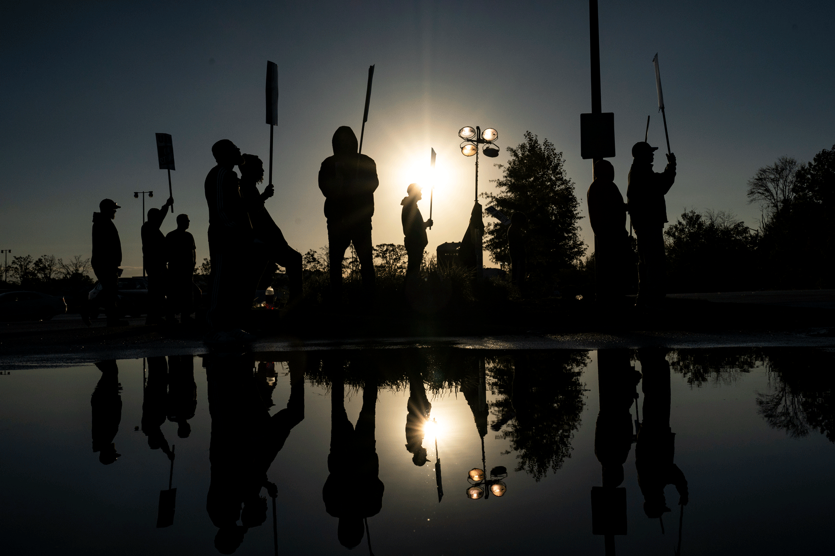 A group of people is seen holding picket signs, with their reflection visible in a puddle in the foreground.
