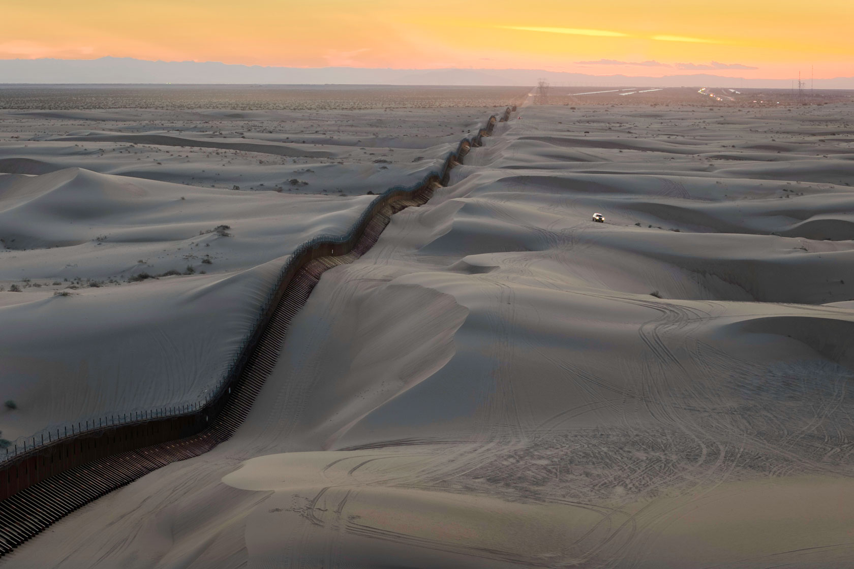 The U.S.-Mexico border wall is seen stretching through sand dunes while the sun sets in the background.