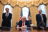 President Donald Trump is seen in the Oval Office signing an executive order, with Secretary of Treasury Scott Bessent and Secretary of Commerce nominee Howard Lutnick on either side of him.