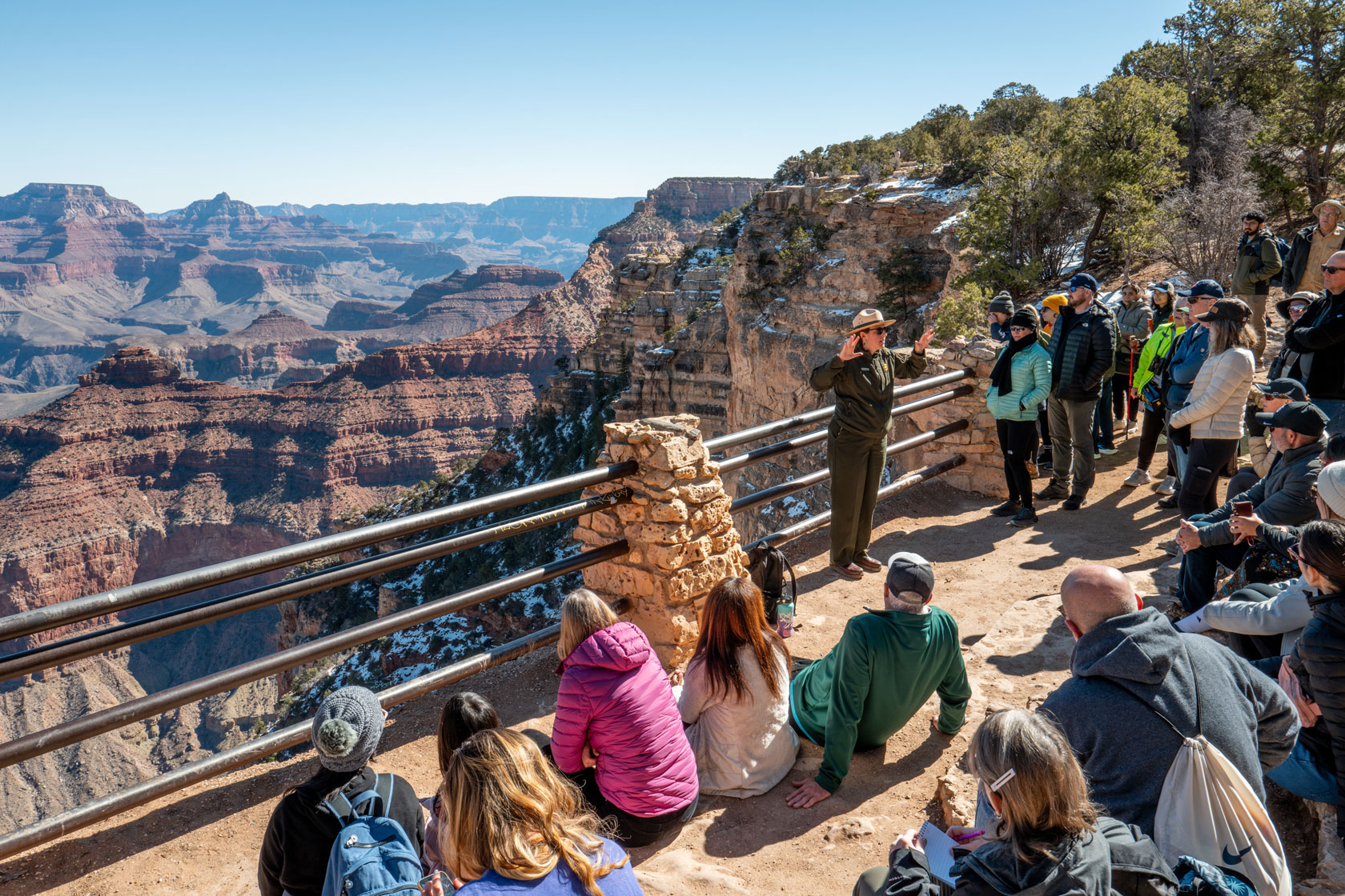A group of visitors is seen receiving a tour from a park ranger at an overlook of the Grand Canyon.