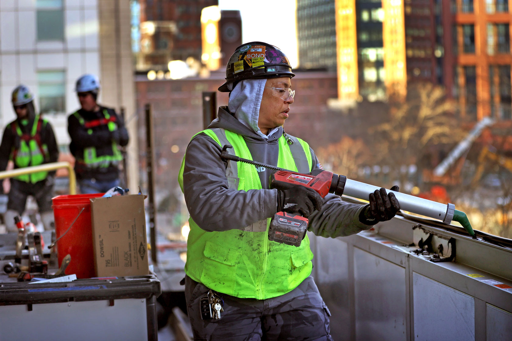 A construction worker is seen using a tool on the side of a building, while two others work behind her.