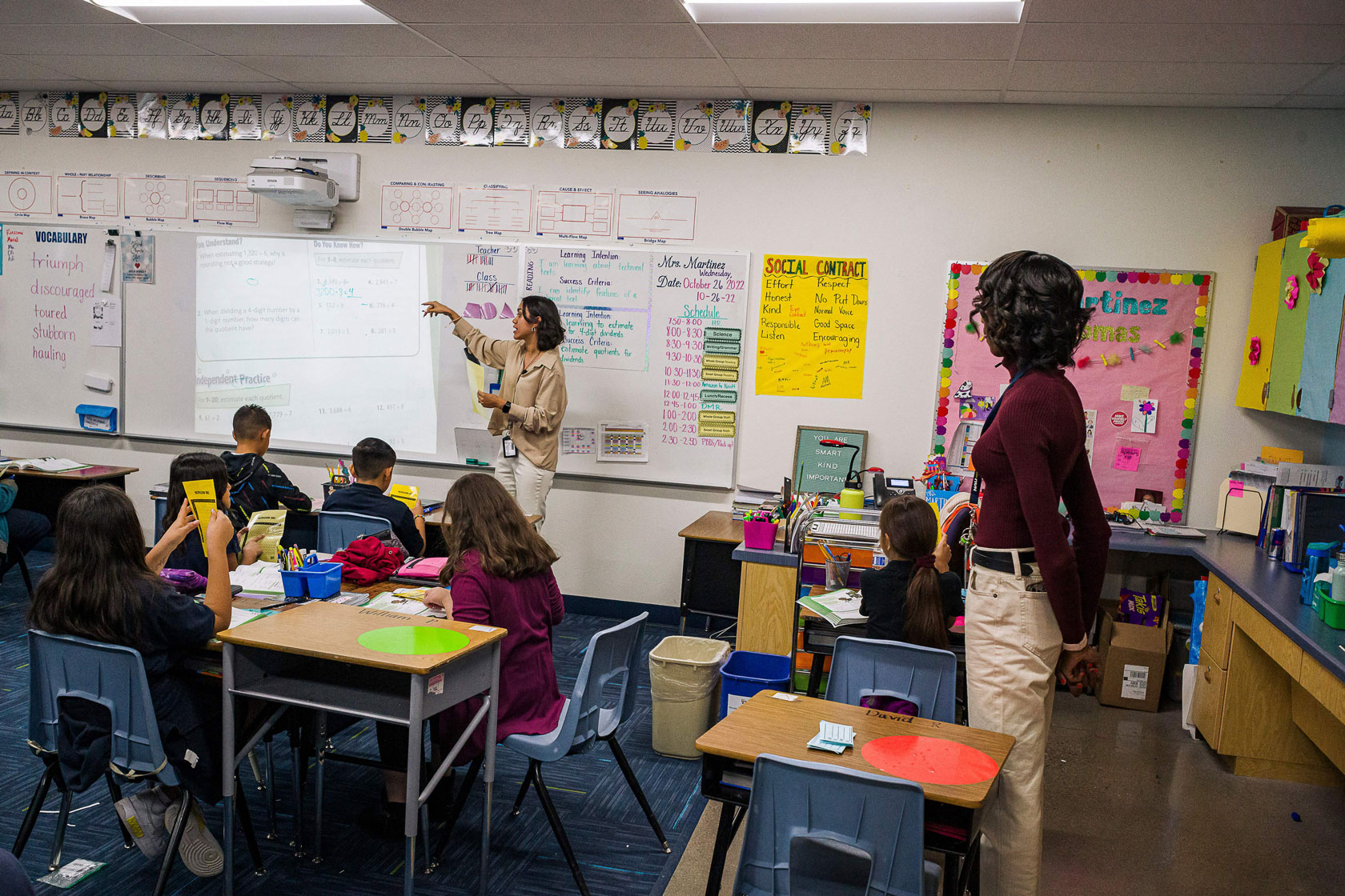 A teacher is seen in a classroom explaining something at the board while students sit at their desks and another teacher watches.