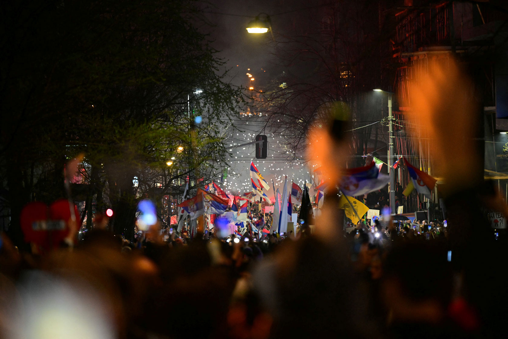 Demonstrators fill a street at night holding Serbian flags and lights.