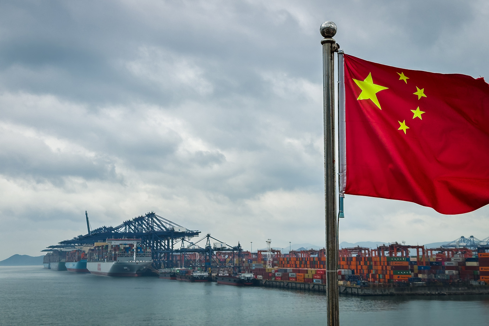 A Chinese national flag flies in front of container ships, cranes, and stacked shipping containers.