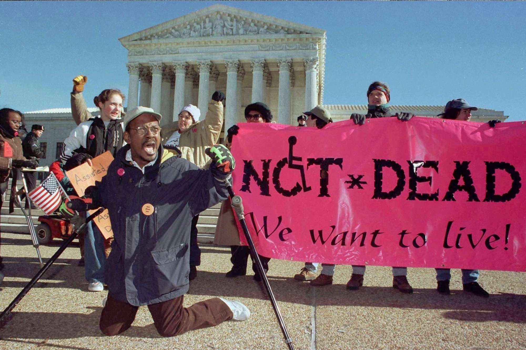 An image of a Gregory Dugan on his knees at a Not Dead Yet protest. A pink banner is held behind him that reads 