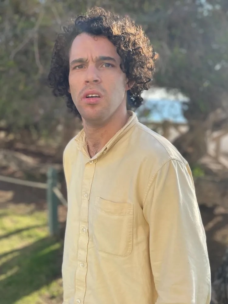 An outside headshot of Reid, a white man with curly hair. He stands outside in a yellow button-up shirt. The sun slightly flares into the shade of surrounding green trees.
