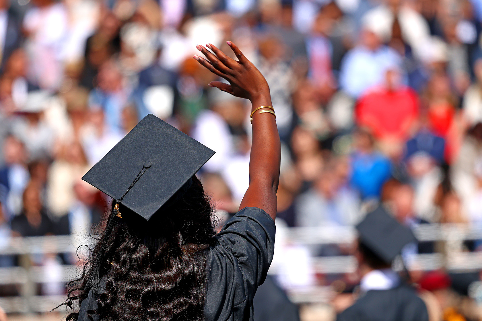 Photo of the back of a graduate wearing a black cap and gown, waving to the graduation audience.