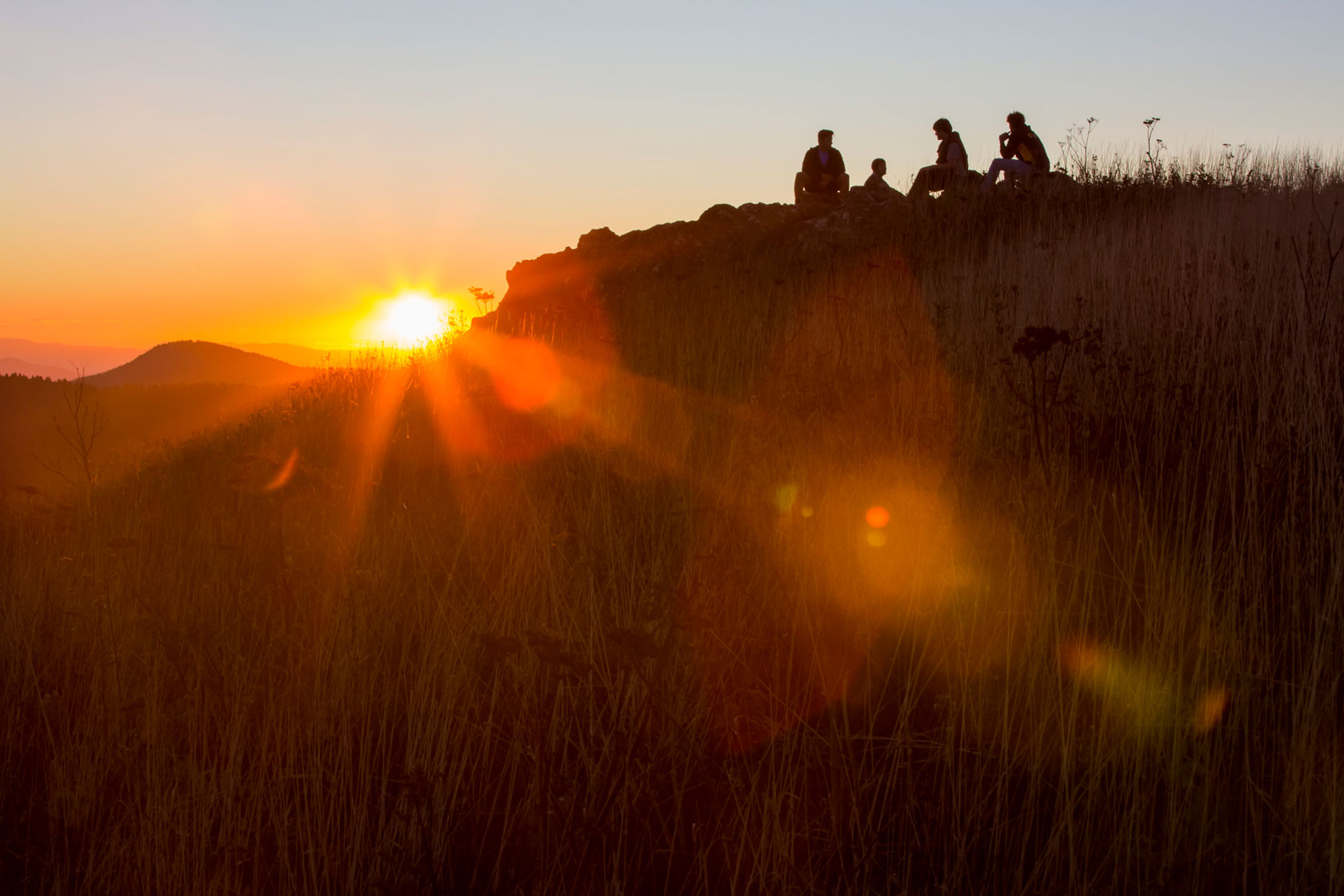 Four people sit atop a ridge as the sun sets in the distance.