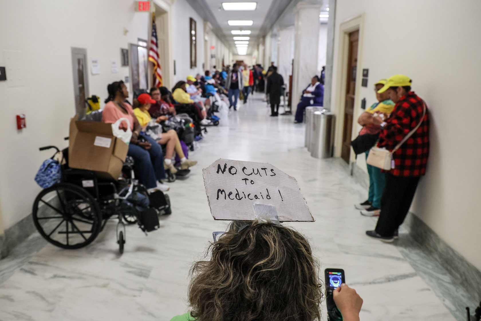 A woman in a wheelchair wearing a sign protesting cuts to Medicaid is seen in a hallway full of protestors.