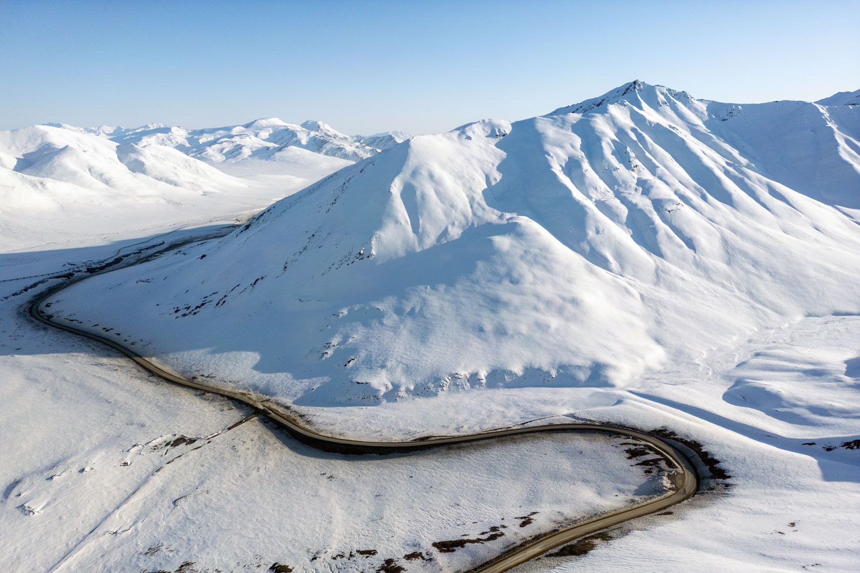 An aerial view of Dalton Highway in North Slope Borough, Alaska, as it curves past a snow-covered mountain range.