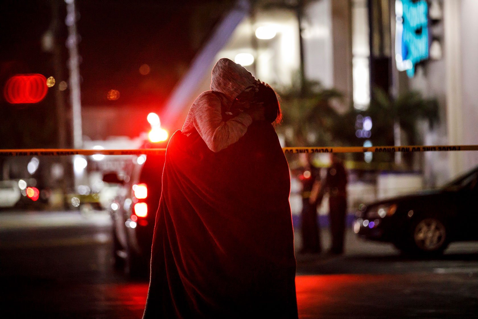 People hug each other in front of a dark crime scene, with the red light from a police car appearing in the background.
