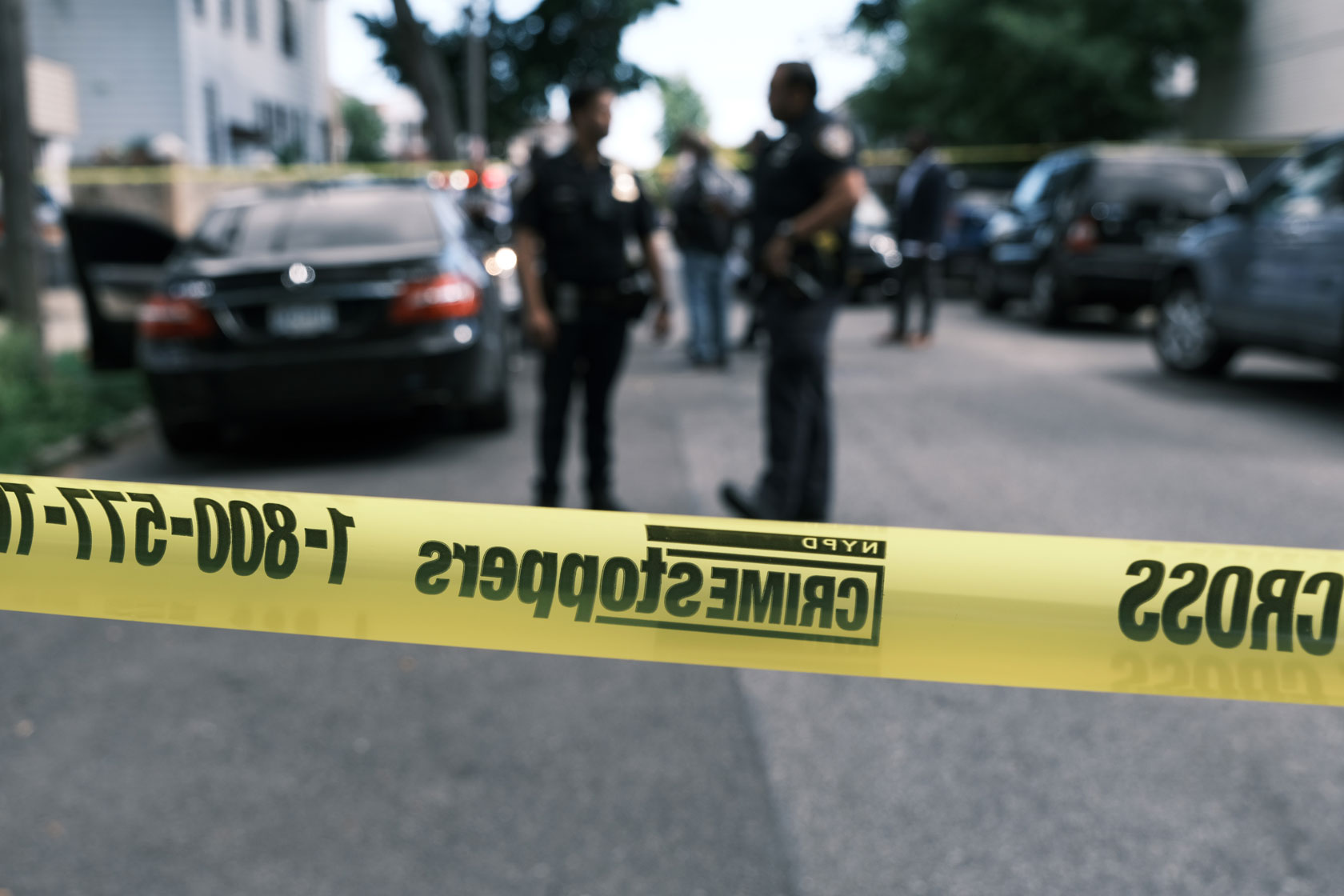 Police officers are seen behind a yellow line of caution tape on a street with cars parked on either side.