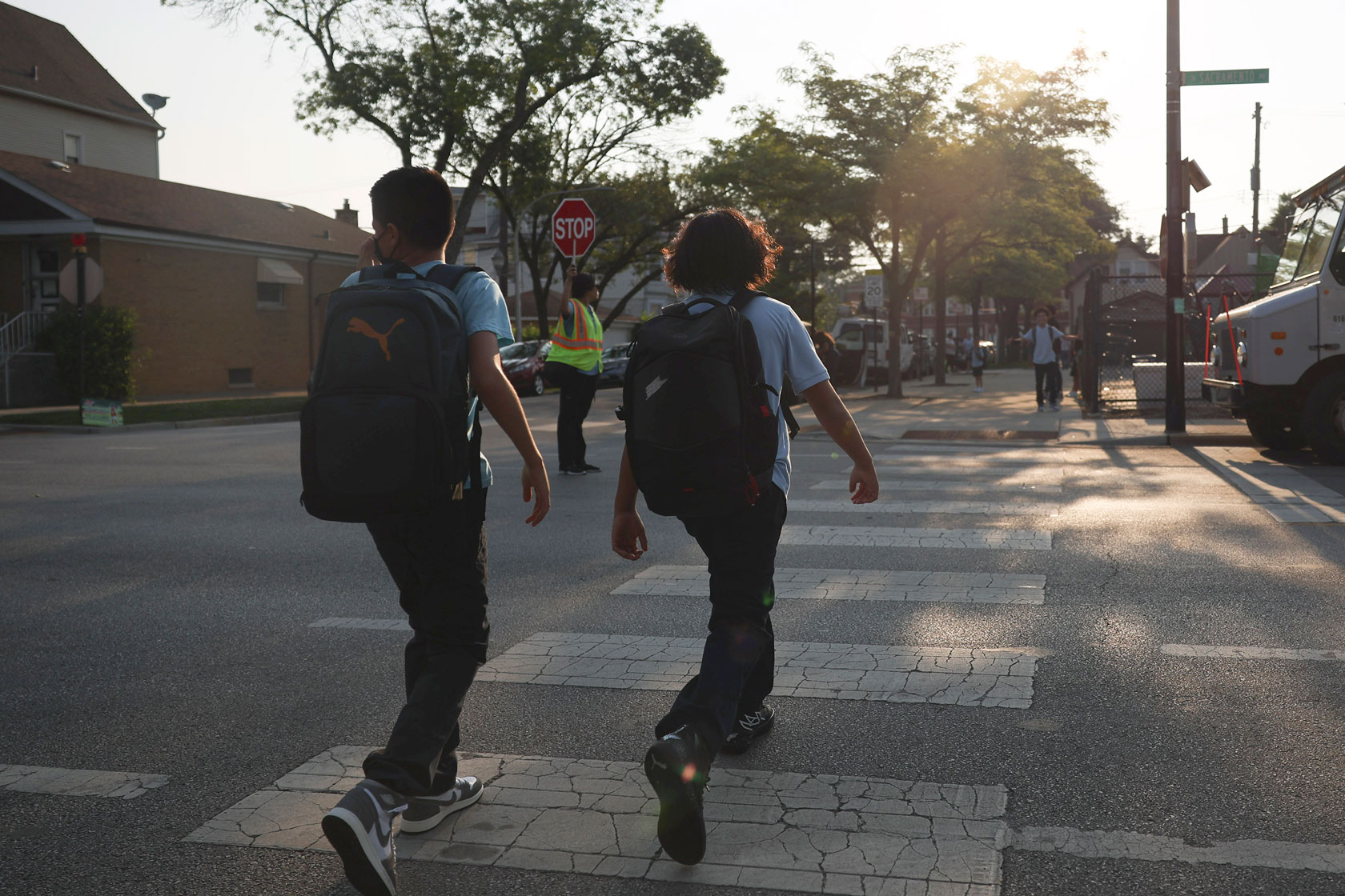 Two students crossing the street wearing backpacks. A crossing guard is in the middle of the street holding a stop sign.