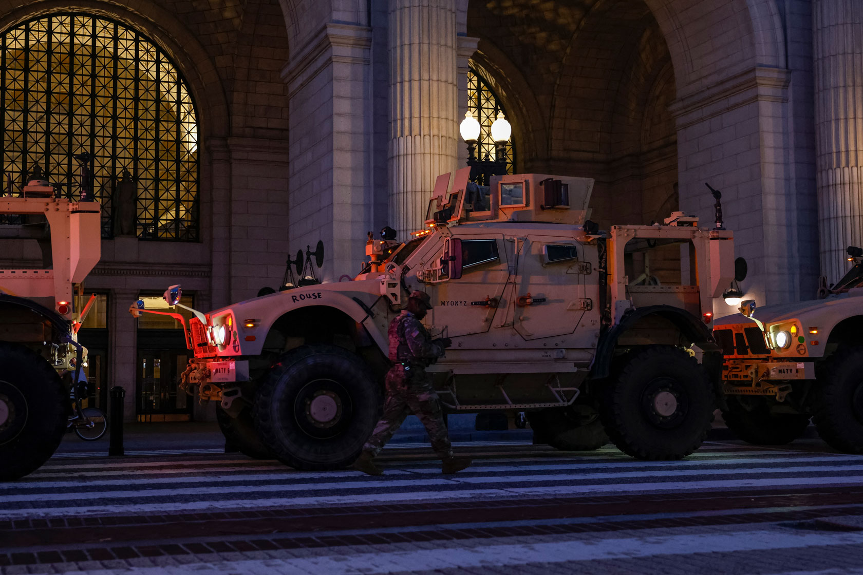 Headlights from military vehicles and a lamp outside Union Station illuminate the street that a member of the National Guard walks along.