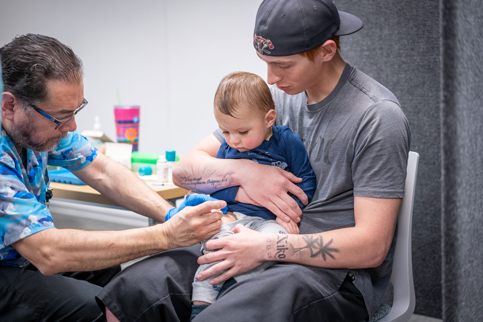 A health care provider vaccinates a toddler in the leg as the toddler and his father look on.