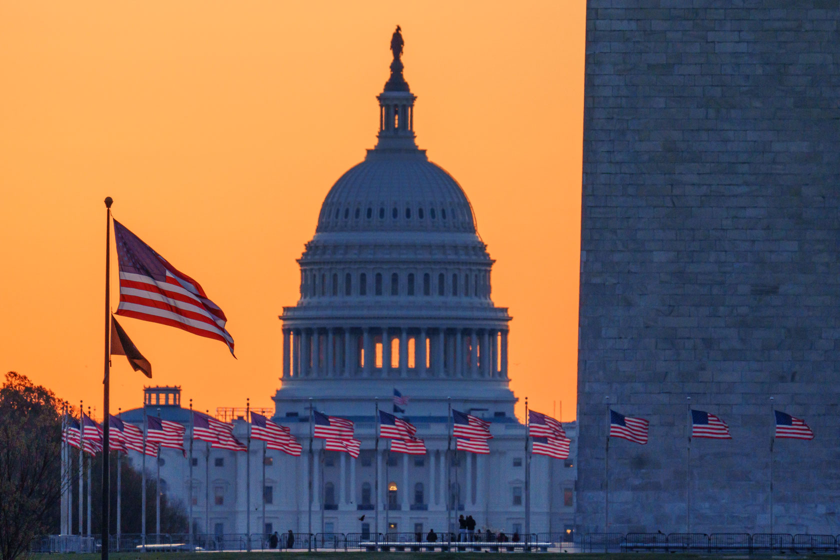 American flags around the base of the Washington Monument dot the foreground as the U.S. Capitol is partially illuminated by the sunrise.
