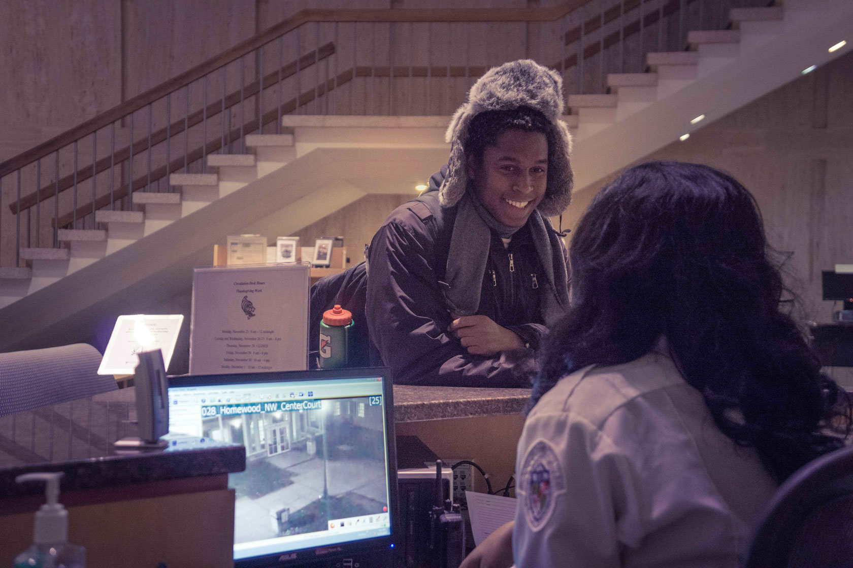 A college student leans on a counter while talking to a security guard seated on the other side.