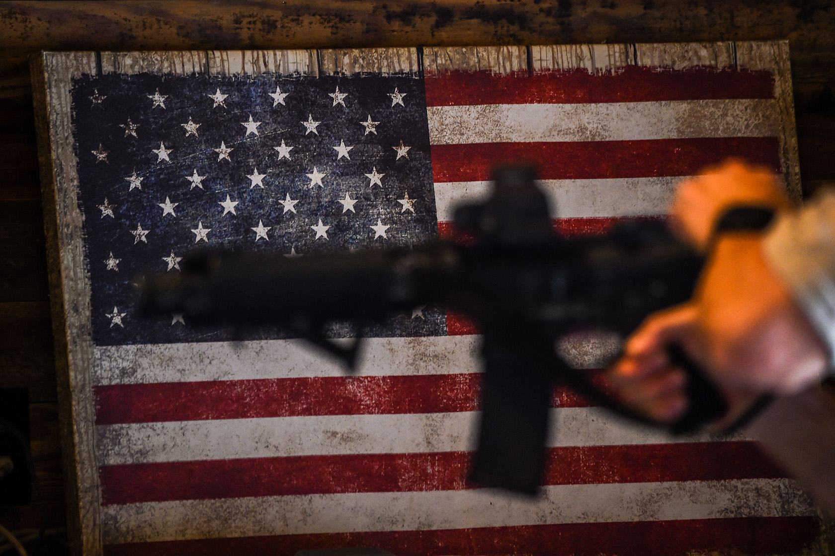 An AR-15 semi-automatic rifle is held in front of a U.S. flag.