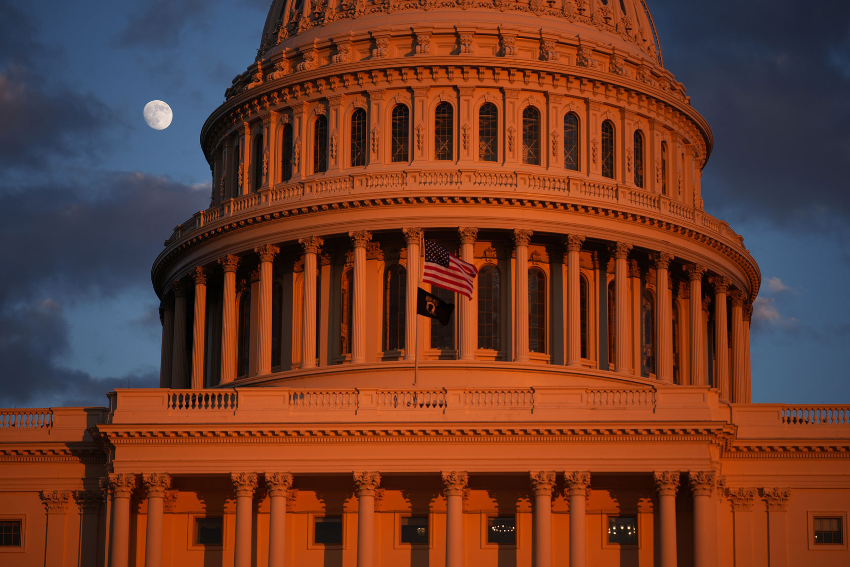 The moon is visible to the left of the Capitol dome in the evening sky.