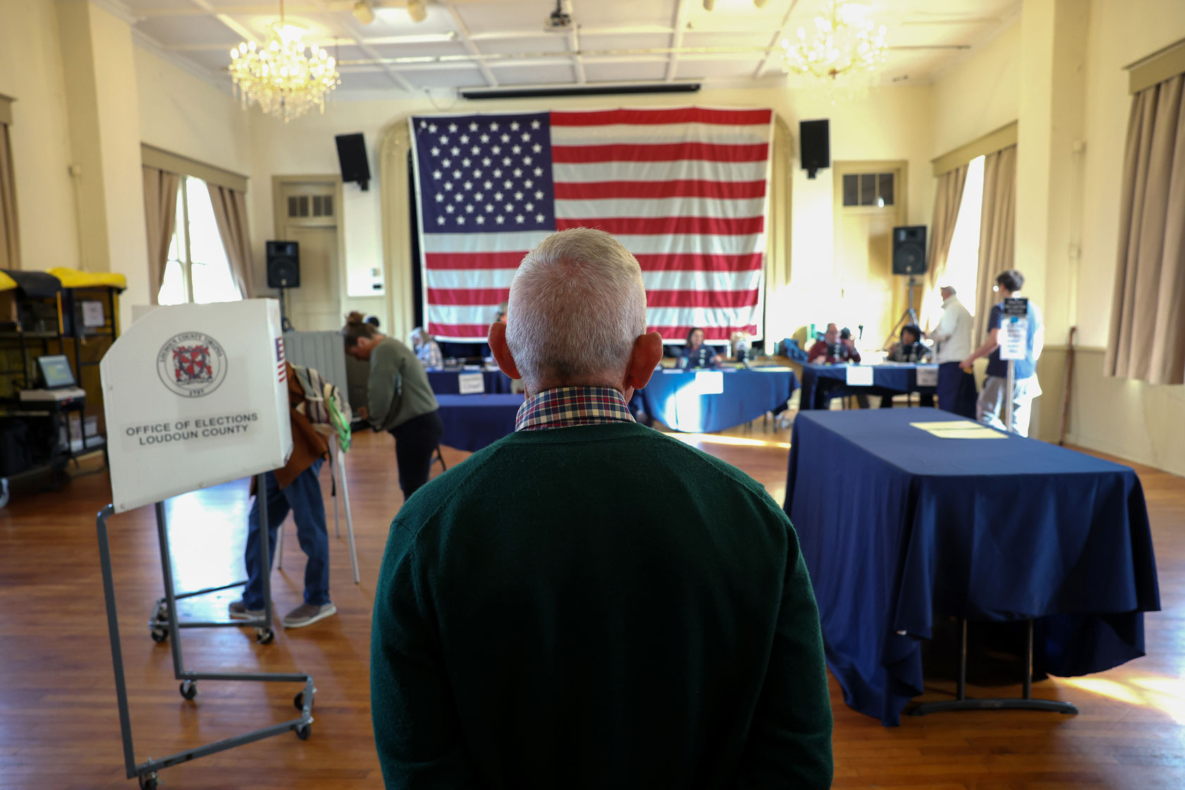 An elections officer looks out over a room where voting booths are set up. An American flag hangs in the background.