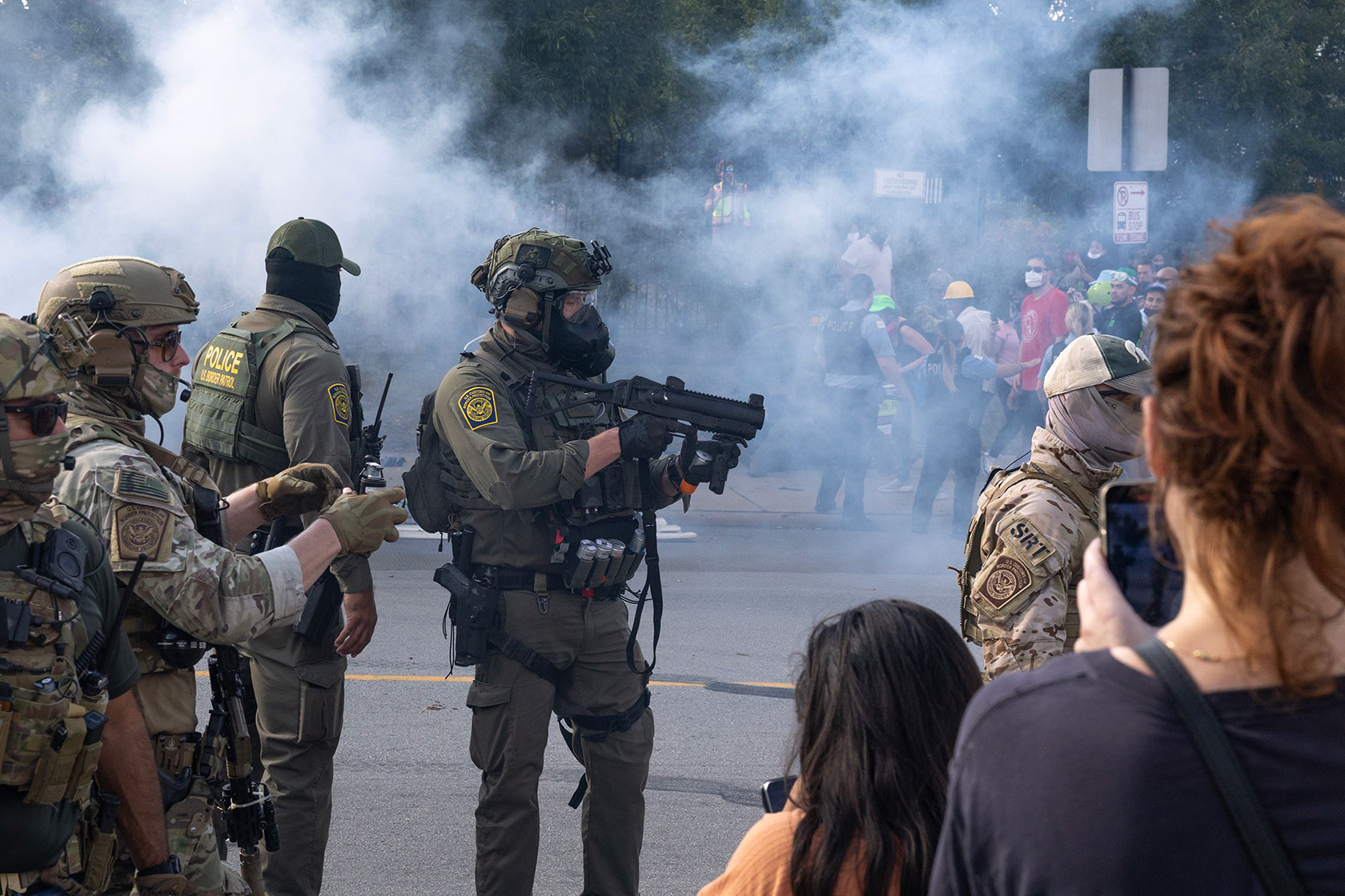 Federal agents, one with gun drawn, walk through tear gas as community members watch; one, in foreground, films on phone