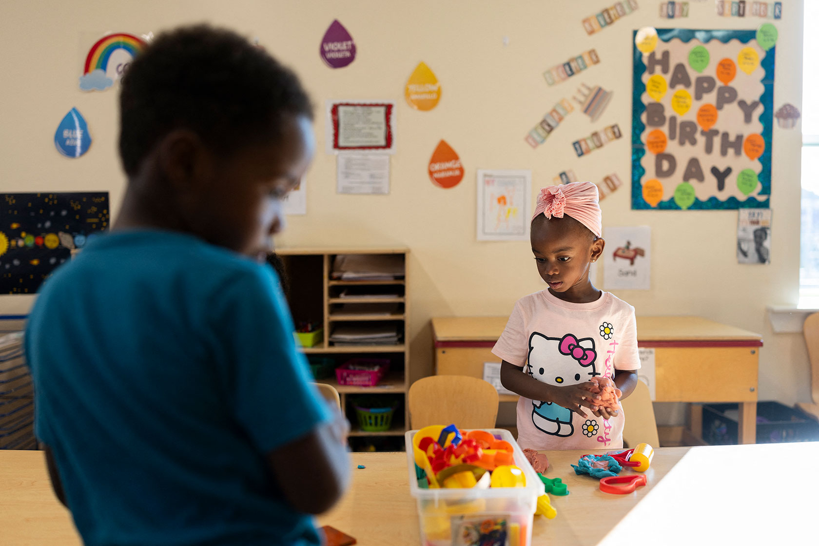 Two young children standing at table, with bulletin board and other decorations in background