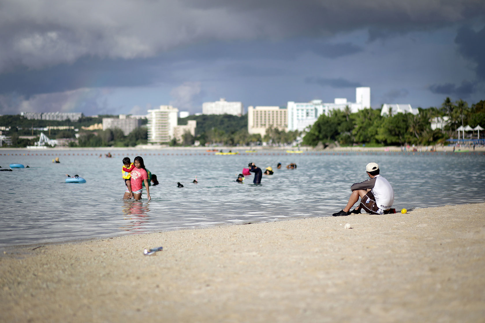 Person sitting on beach and others in the ocean, with city buildings in the background