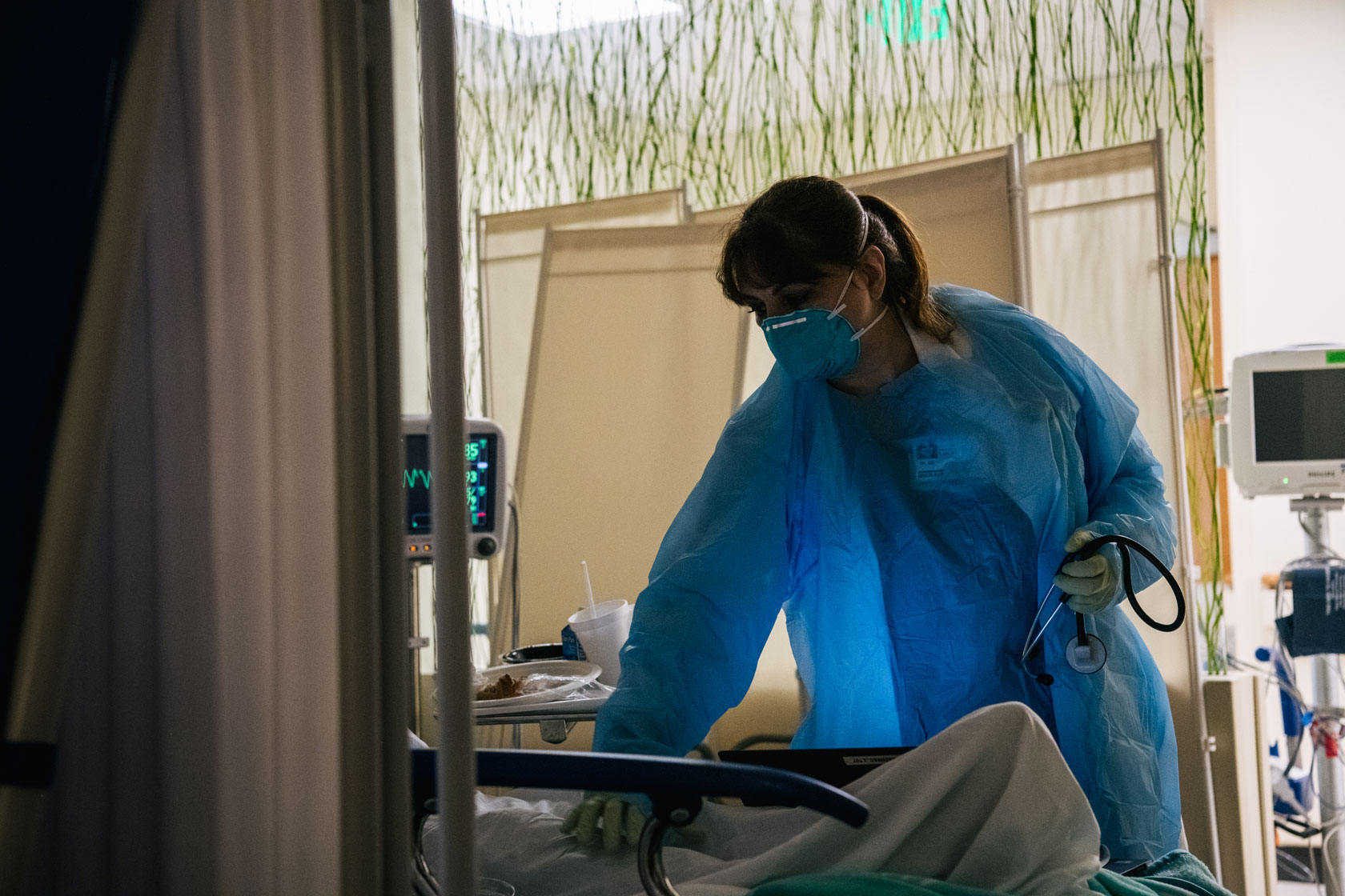 A masked doctor in a blue gown leans forward to tend to a patient lying in a hospital bed.