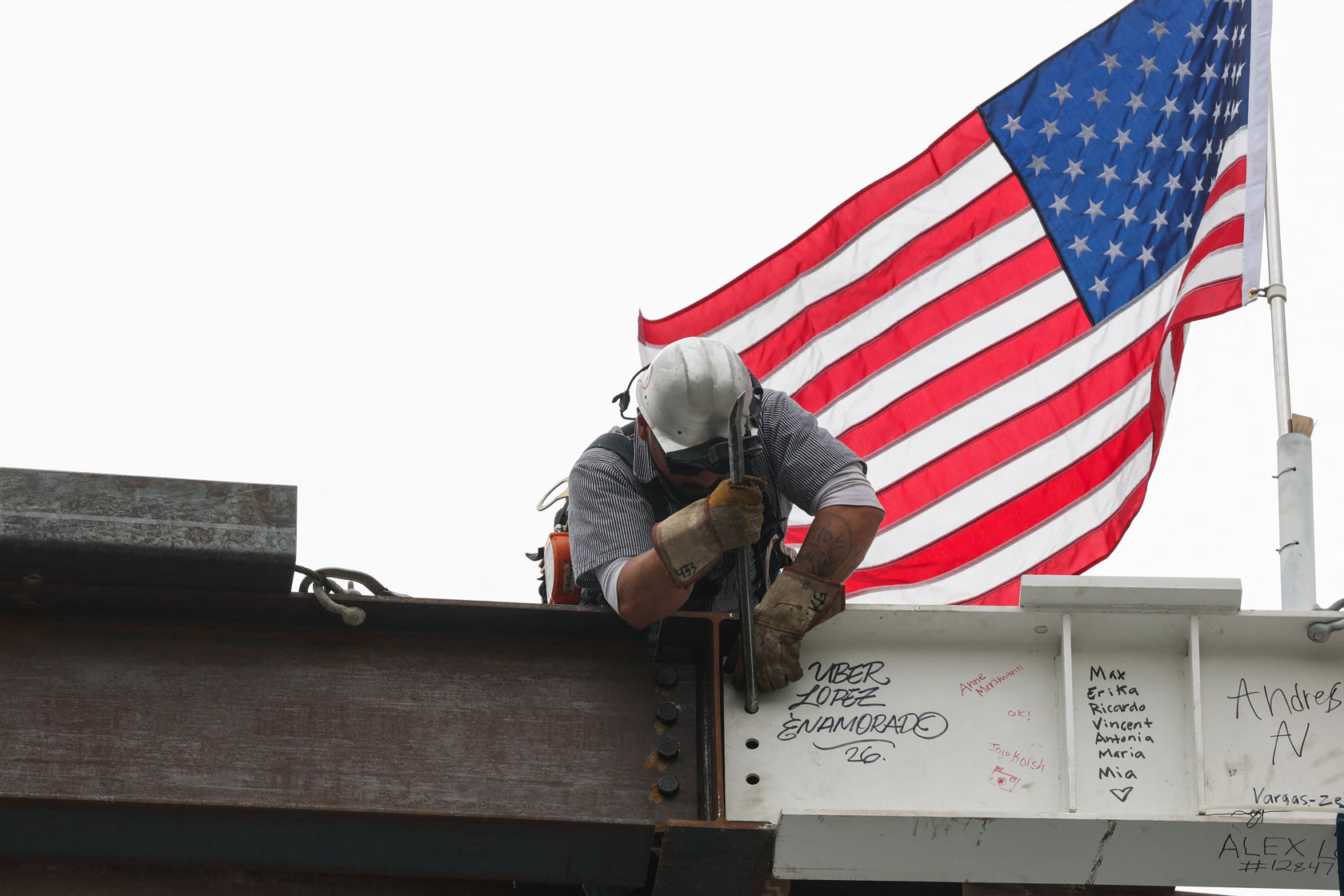 A steelworker leans forward as he installs a beam. An American flag flies from a pole attached to the beam.