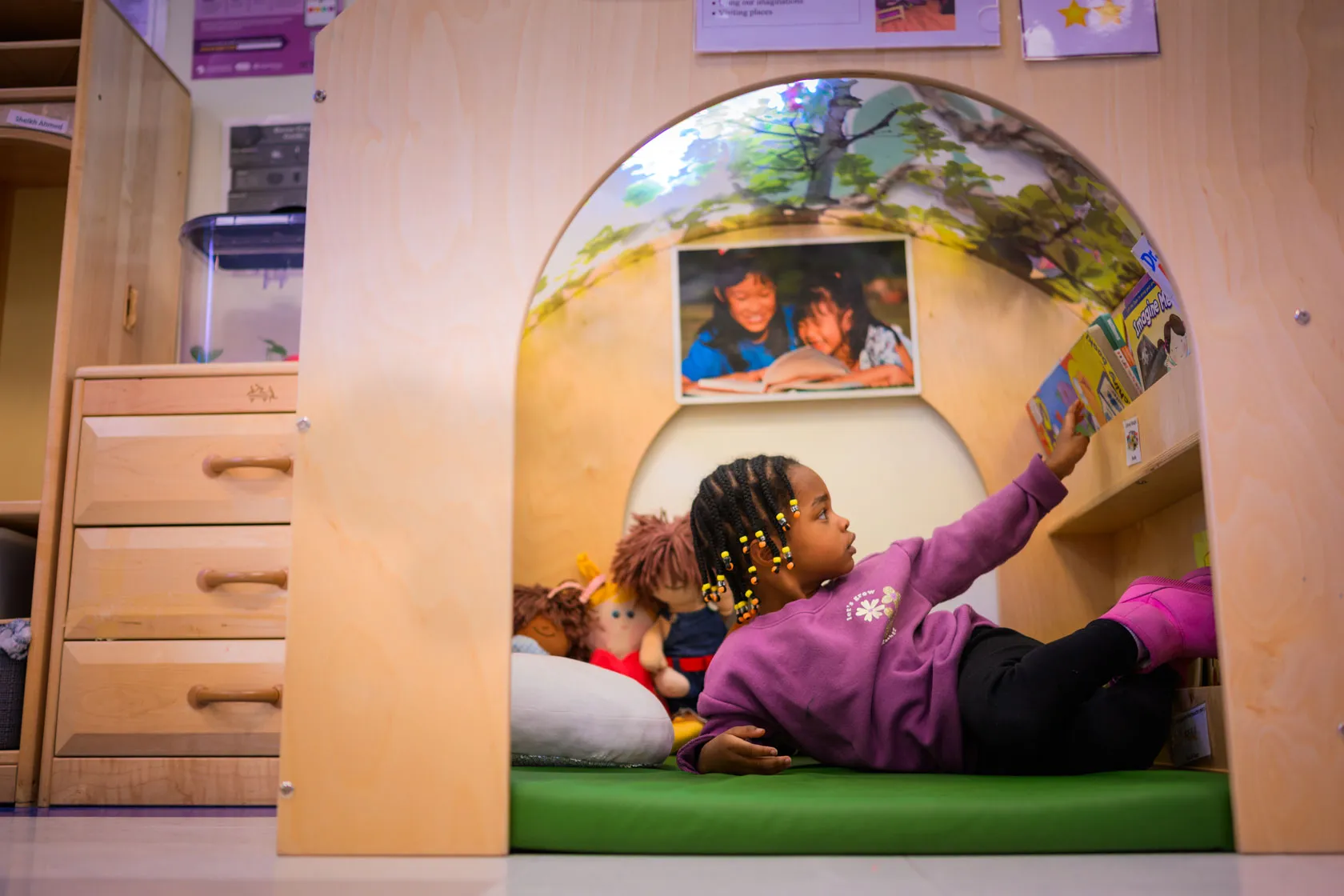 A child lying on a mat reaches for a book on the shelf.