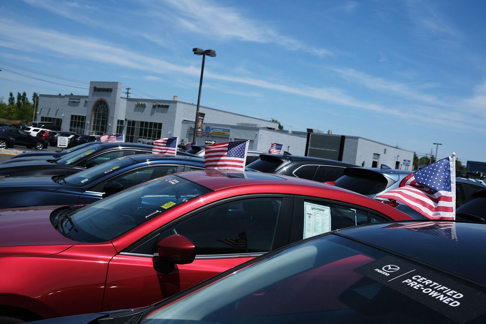 A row of cars for sale at a dealership, each flying a small American flag from its roof, parked in front of the dealership building under a mostly clear blue sky.