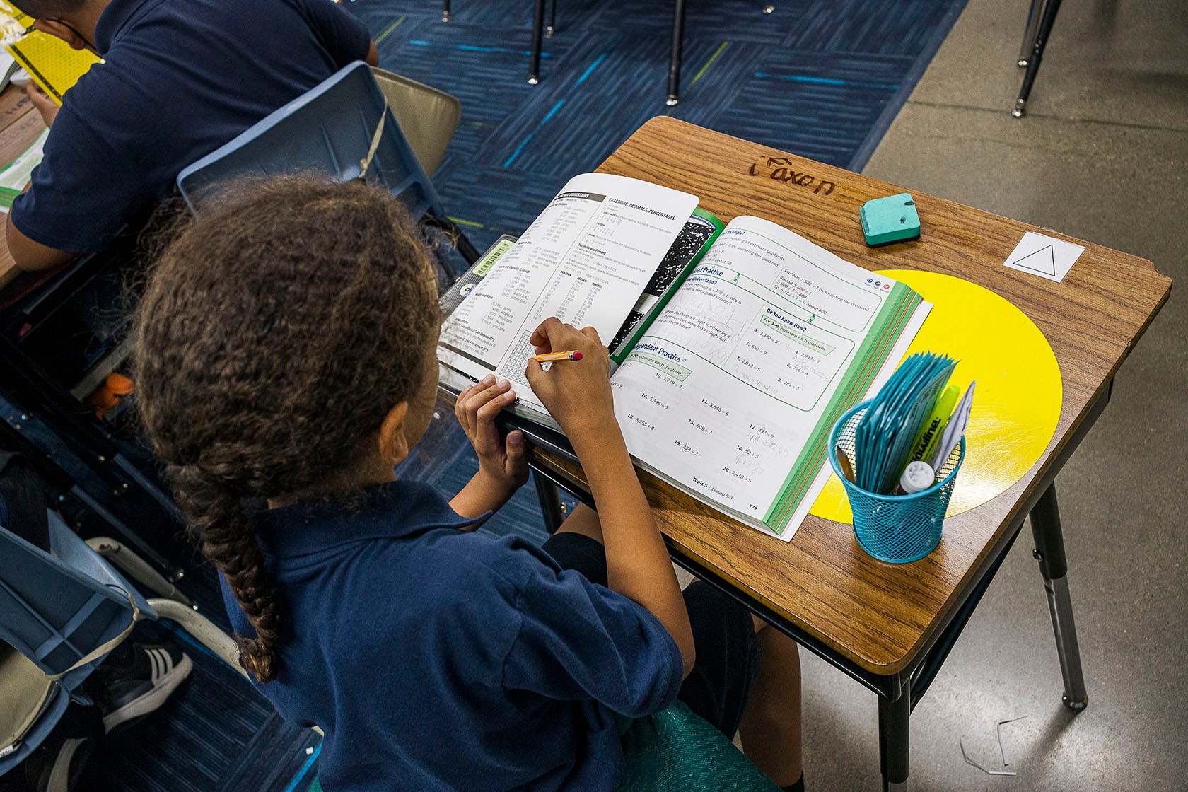 A student sitting at a desk writes in an open math workbook.