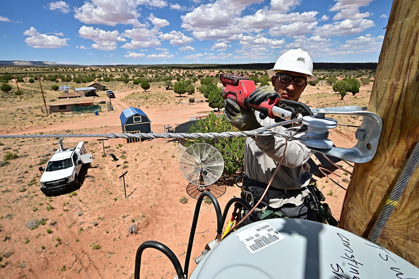 A utility line worker in a hard hat and safety glasses uses a hydraulic tool to secure a cable to an insulator on a wooden pole, with a rural property of small buildings and a satellite dish visible below under a partly cloudy sky.