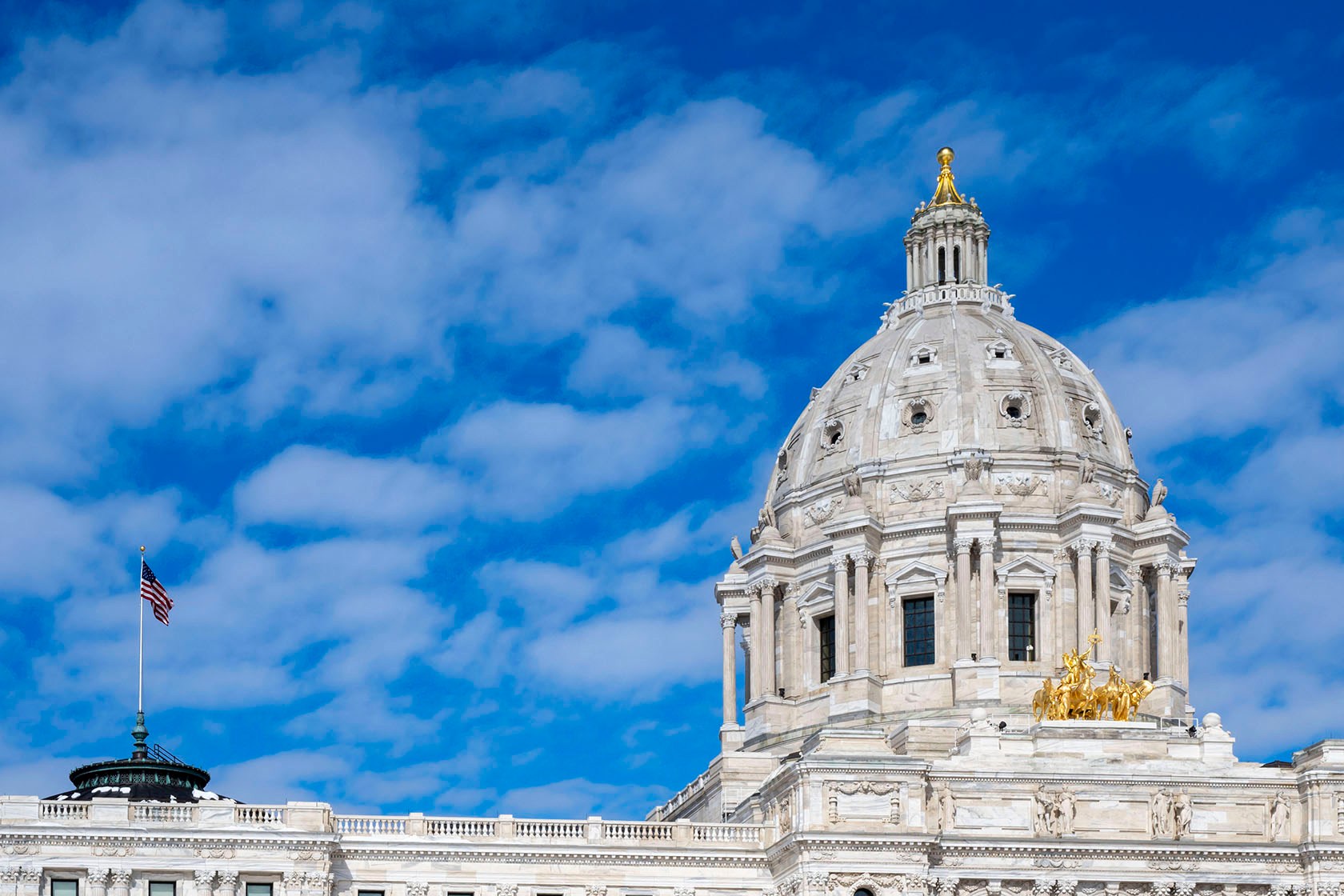 The dome of the Minnesota State Capitol is framed by a blue sky and clouds. An American flag flies to the left of the dome.