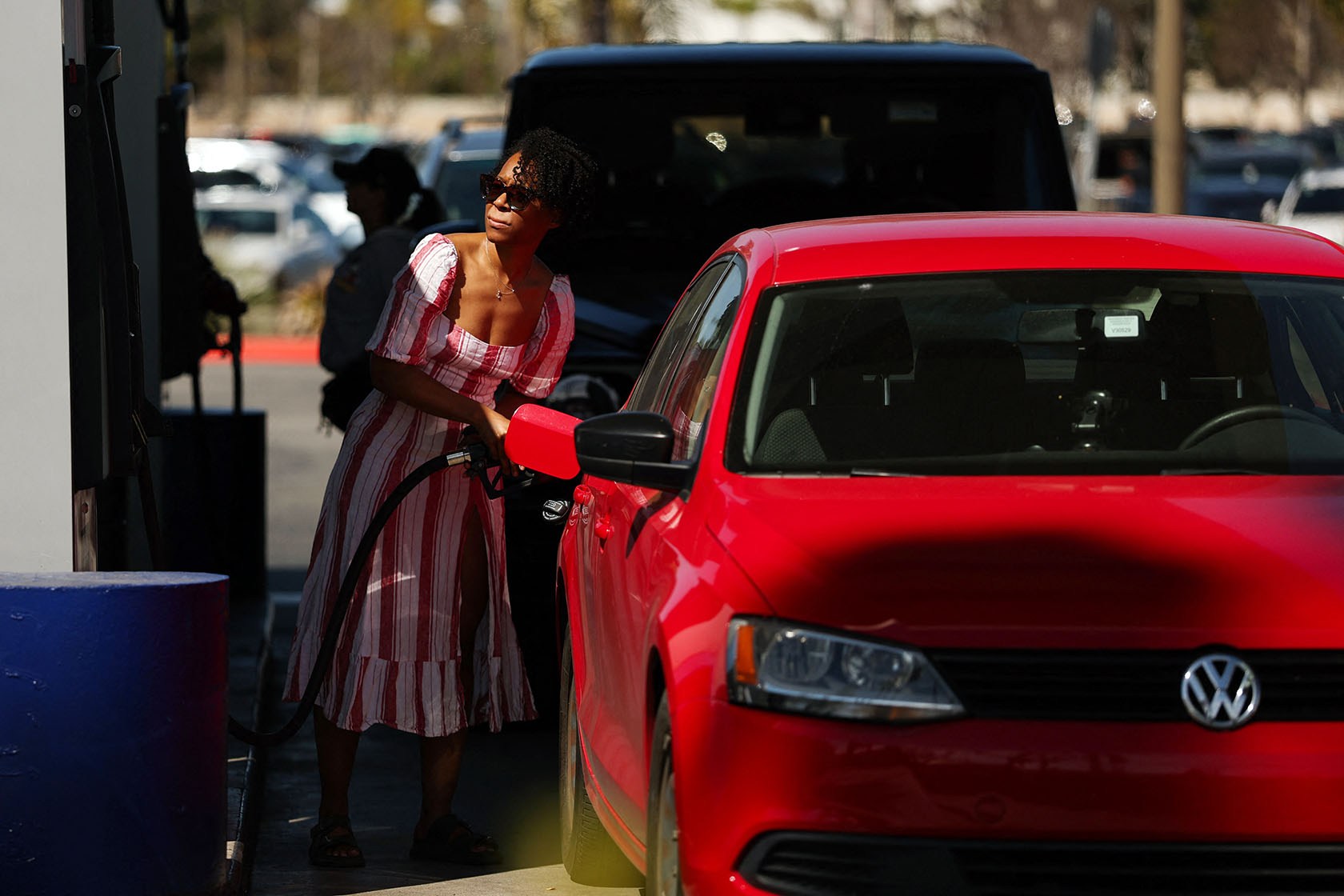 A woman wearing a red-striped sundress fills up a red sedan at a gas station pump on a sunny day.