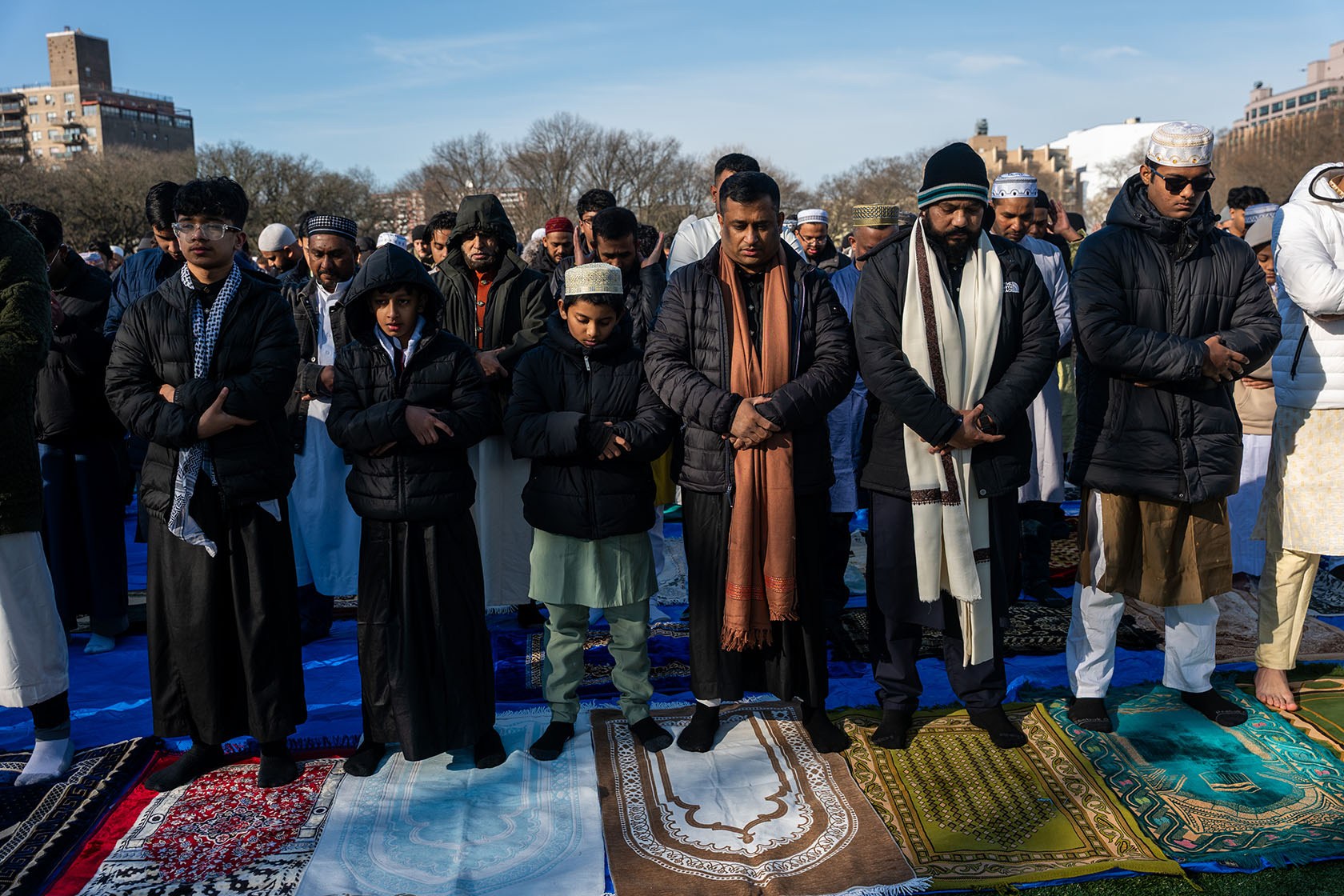 Worshippers with folded arms and bowed heads stand shoulder to shoulder on prayer rugs laid over the ground.