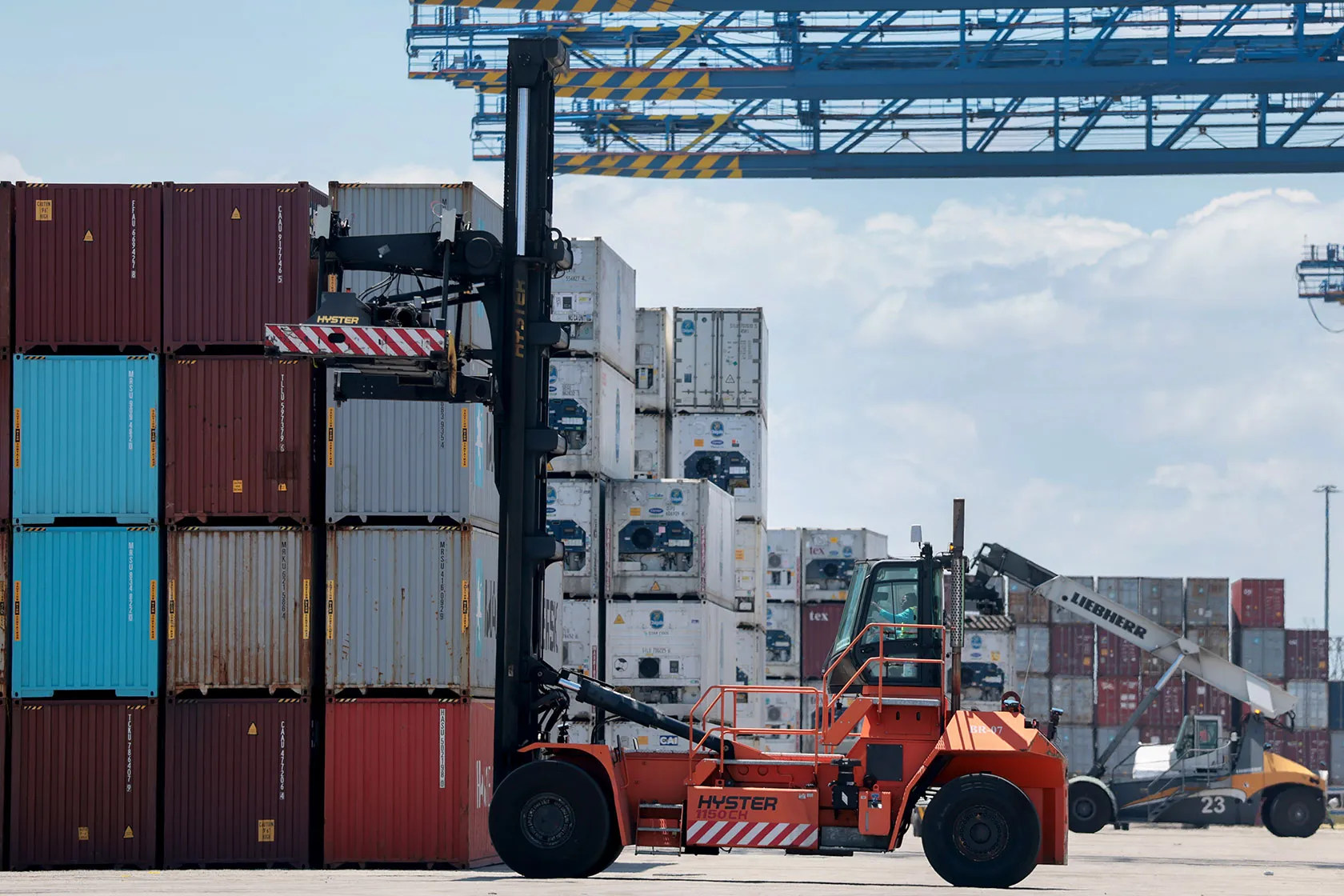An orange container handler with its mast raised beside towering stacks of shipping containers at a port, a crane overhead