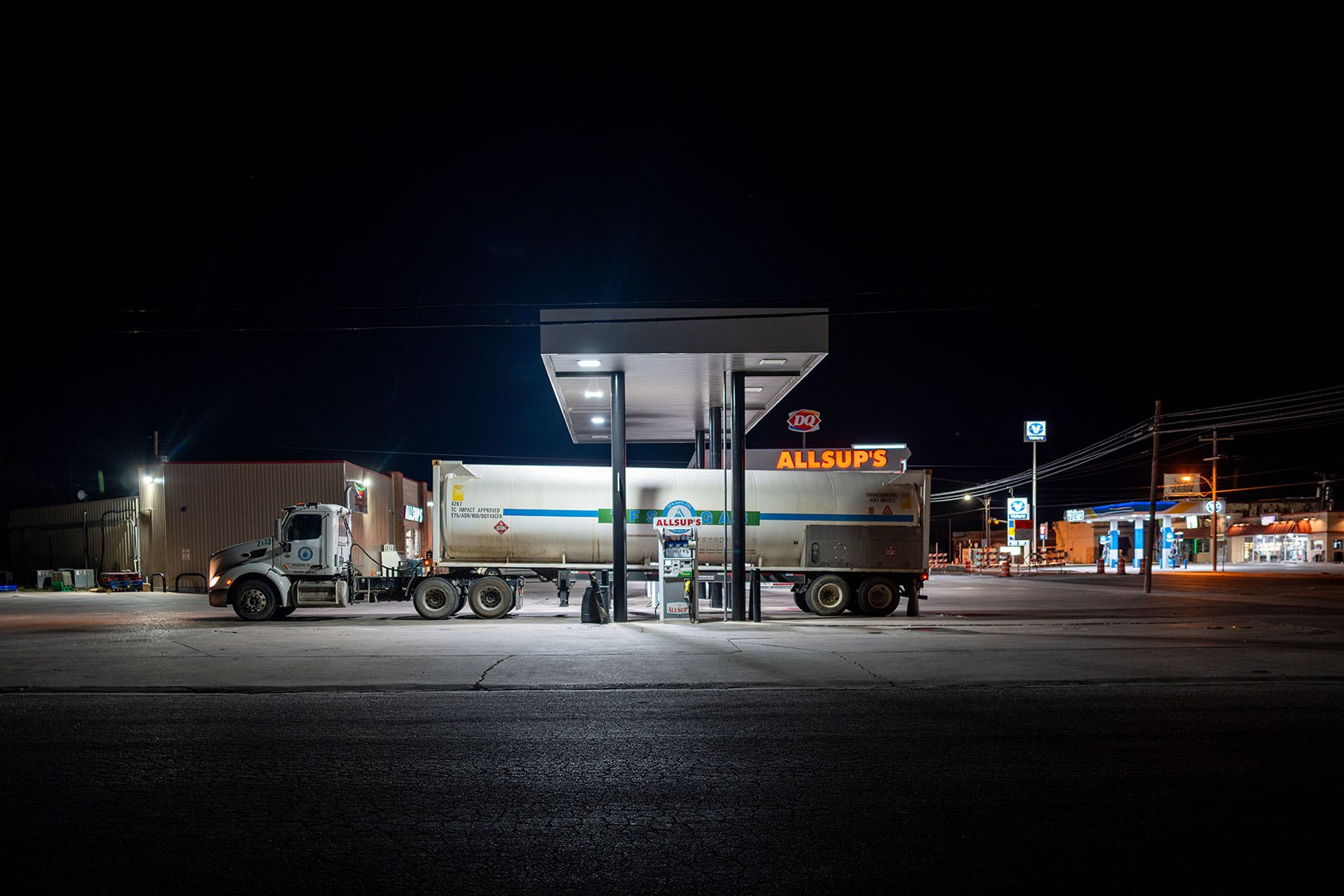 A semi-truck refuels beneath the lit canopy of an Allsup's gas station, with a Valero station glowing down the empty road in the distance.
