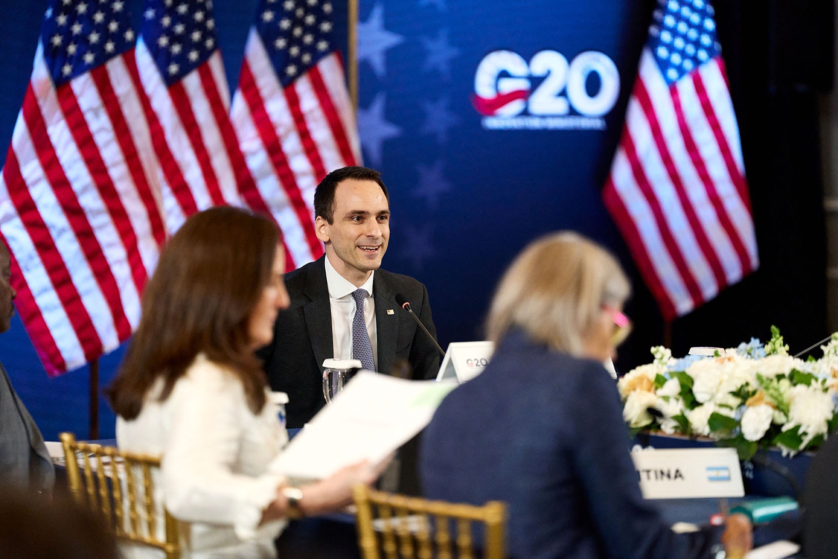 Michael Kratsios sits at a table, with American flags and the G20 logo behind him in the background.