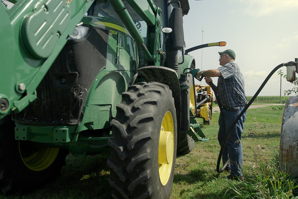 A farmer fuels a large tractor from a storage tank, with a cornfield in the background.