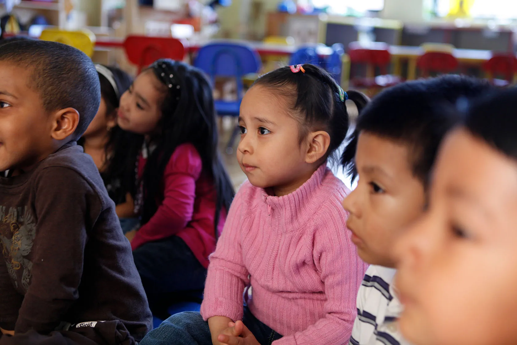 Young children sit together on the floor, their attention focused on something out of frame, in a classroom; small, colorful chairs can be seen in the background.