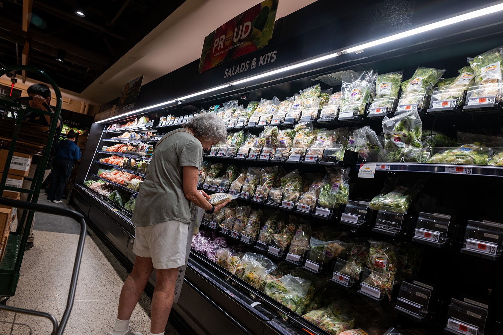 A gray-haired woman looks at a packaged salad kit while standing in front of a refrigerated case.