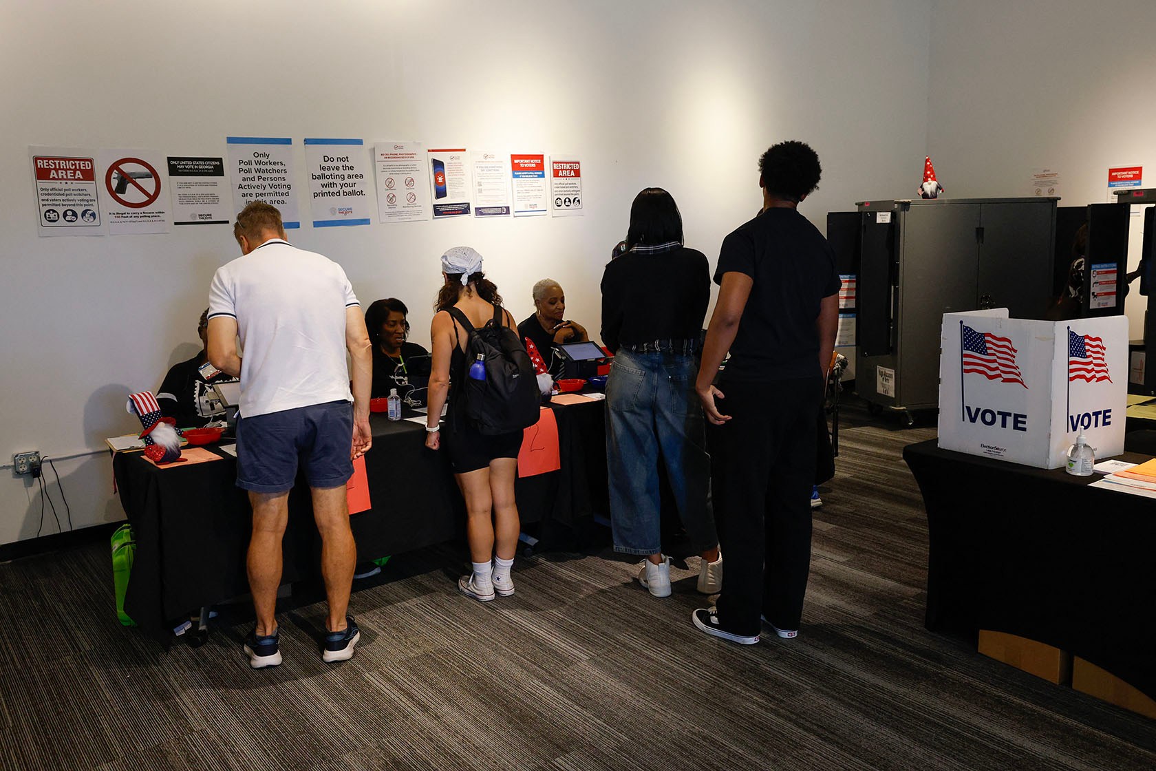 Four voters stand at a check-in table. Above the election workers seated at the table are multiple wall signs, including one that says “Restricted Area” and a sign to tell voters not to leave the balloting area with their printed ballot. White “VOTE” privacy screens and a voting machine are at right.